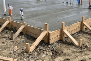 A freshly poured concrete foundation slab with plumbing pipes and wire sleeves protruding through the surface, supported by wooden forms and braces at a residential construction site.